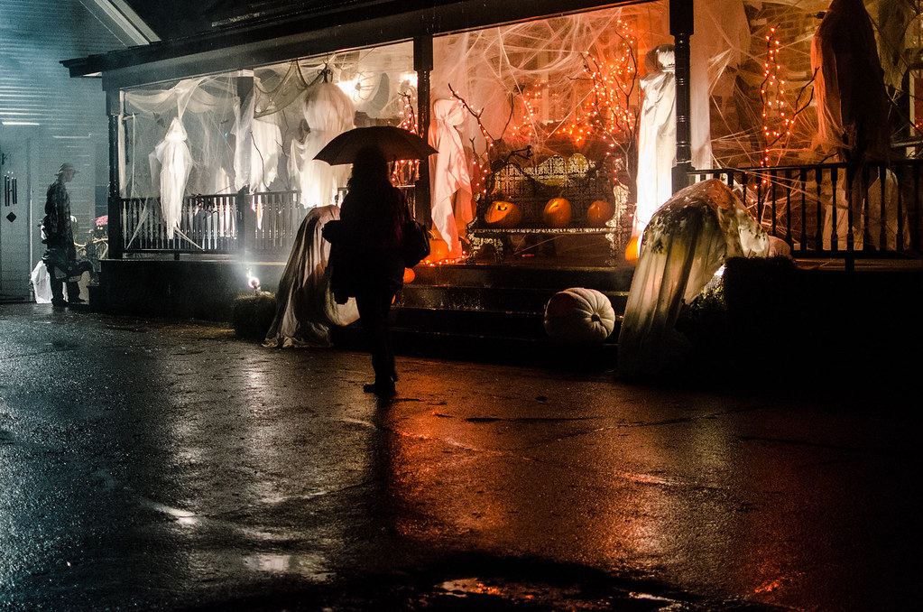 An image of the frontage of a haunted house. Several ghosts are hanging up, as well as netting and cobwebs hanging from the ceiling. There are three pumpkins at the front and orange lights which add to the spooky atmosphere. A woman carrying an umbrella is walking past in front.