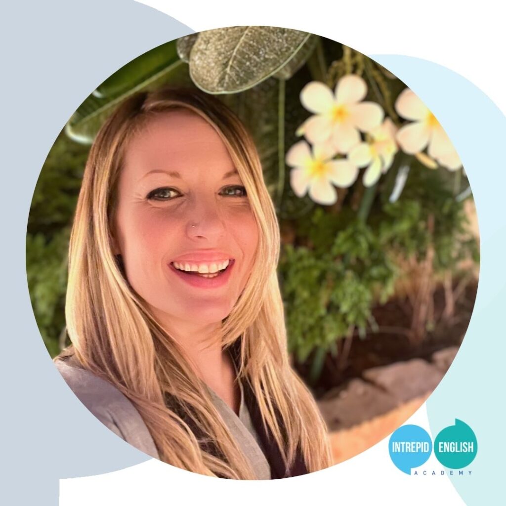 Intrepid English Founder Lorraine is smiling and taking a selfie in front of some beautiful white and yellow flowers. Her blonde hair is down and she is wearing a grey shirt.