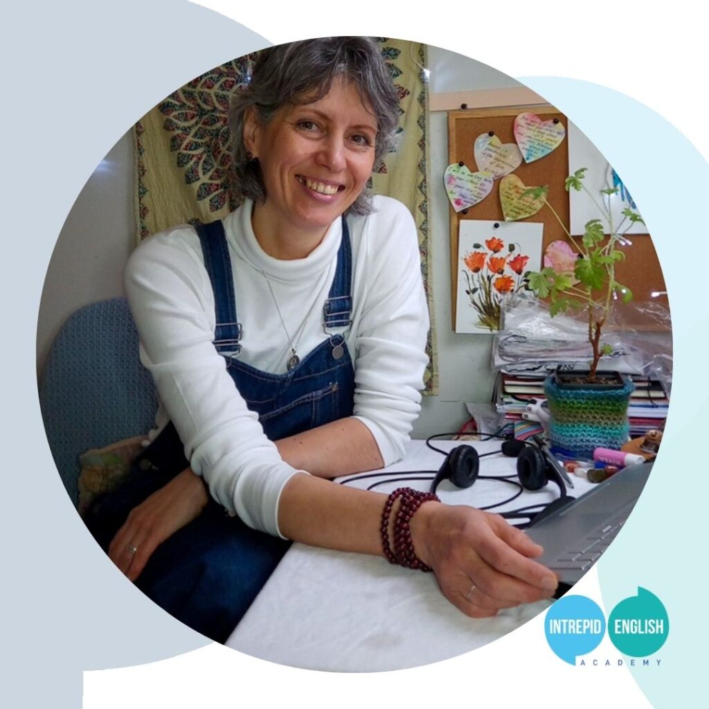 Intrepid English Teacher Natalia is sitting at her desk and smiling at the camera. In the background a pinboard is visible with notes and a picture of some orange flowers. Natalia is wearing a white top and blue dungarees.