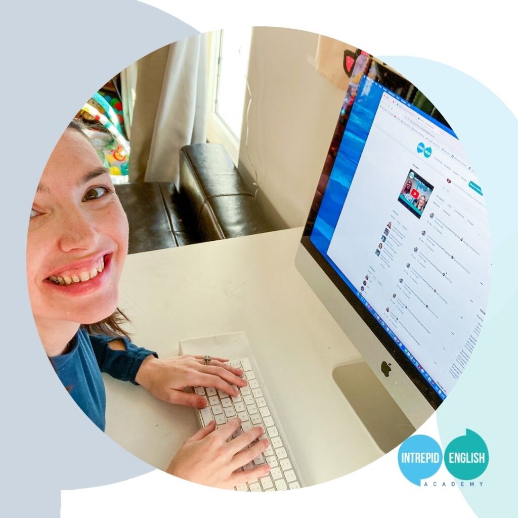 Intrepid English Teacher Kate R is sat at her desk and typing on her keyboard. Her computer is open on the Intrepid English homepage. She is looking up at the camera and smiling. She is wearing a long-sleeved shirt in Intrepid English blue.