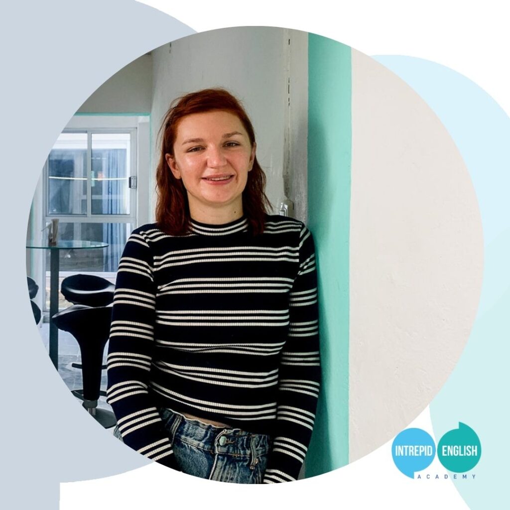 Intrepid English Teacher Kate B is leaning against a white and turquoise wall and smiling at the camera. She is wearing a long-sleeved shirt with dark blue and white stripes.